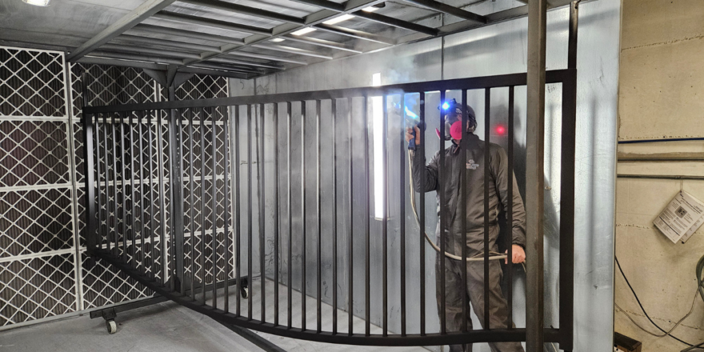 Technician applying powder coating to large metal gate with vertical bars inside spray booth